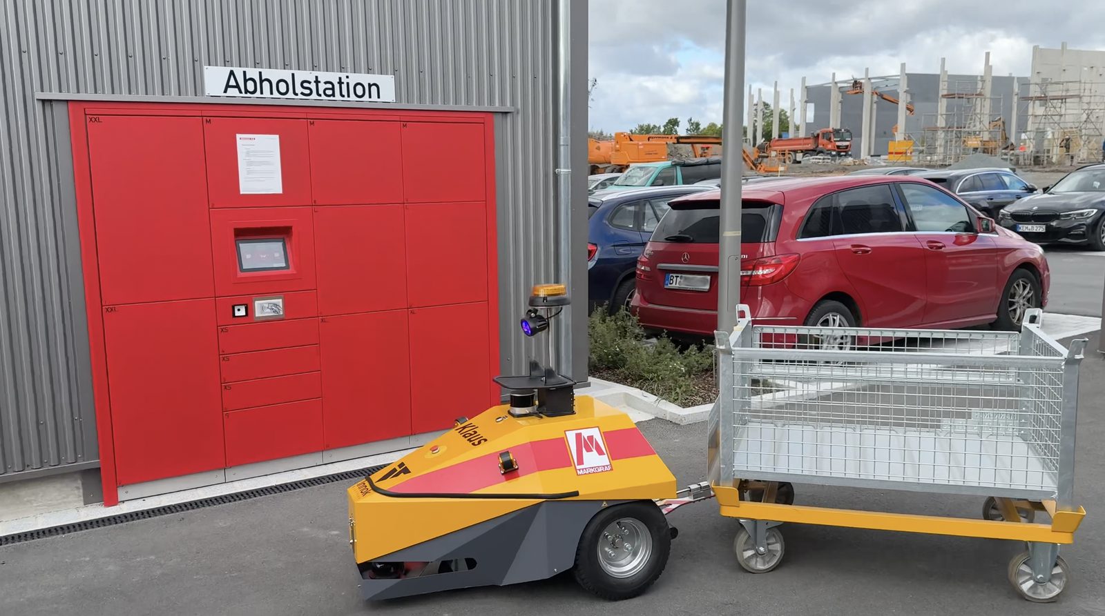 Yellow INDUROS transport robot towing a mesh trolley in front of a parcel pickup station – factory logistics at Markgraf