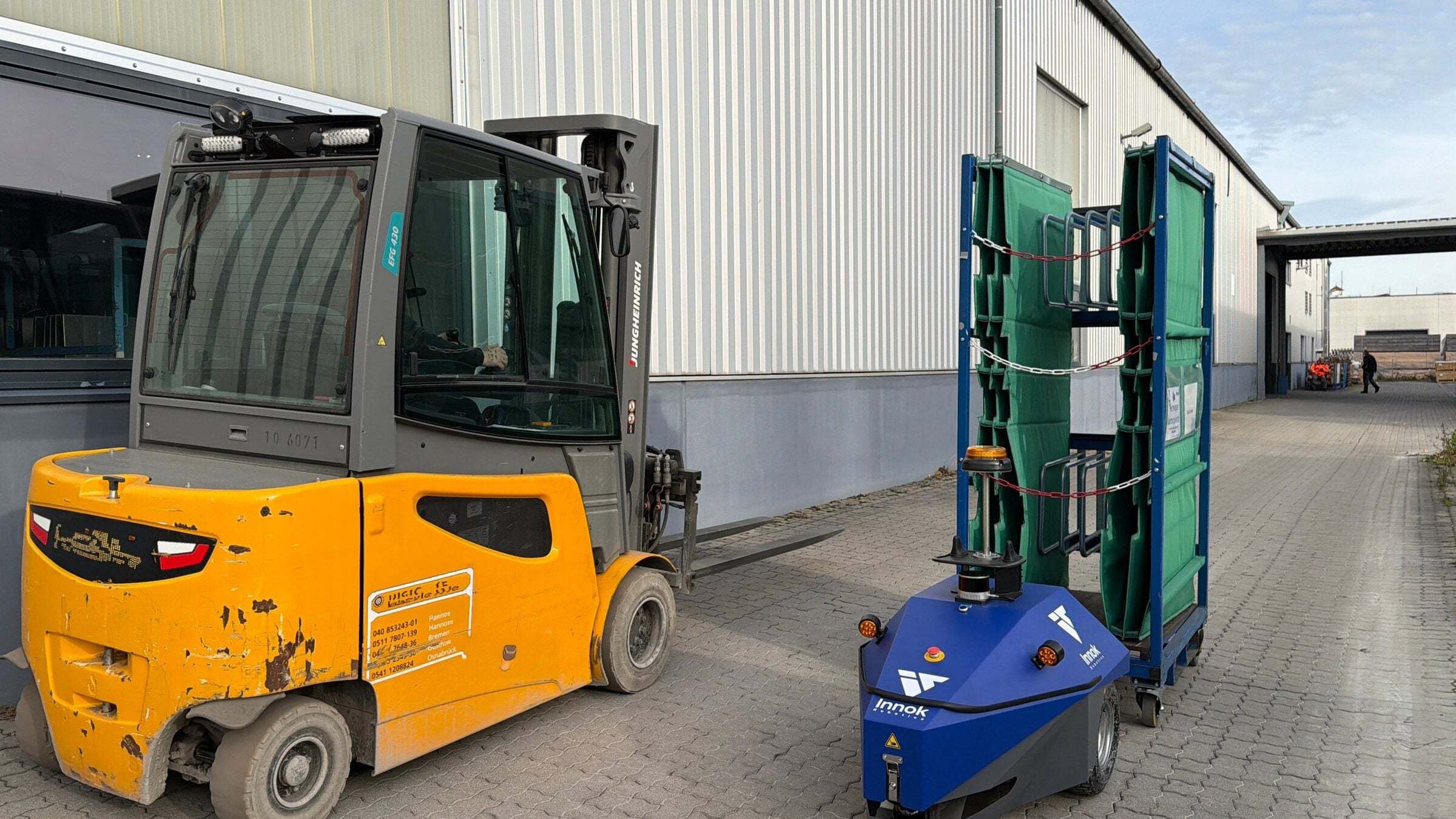 Forklift next to the INDUROS autonomous transport robot with a trailer outside a factory hall