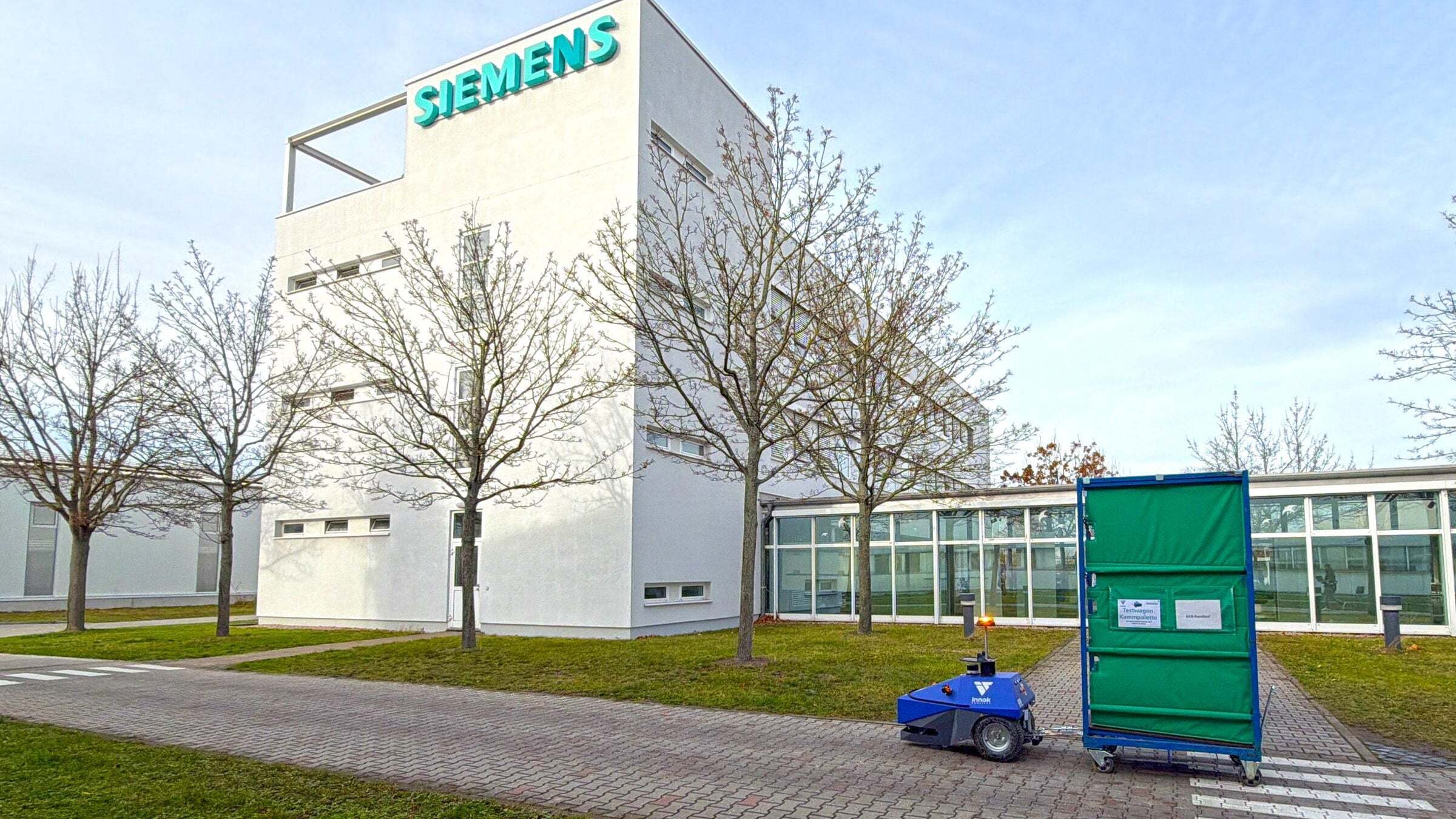 Transportroboter INDUROS zieht einen grünen Rollwagen vor einem Siemens-Gebäude – Integration in Siemens SIMOVE