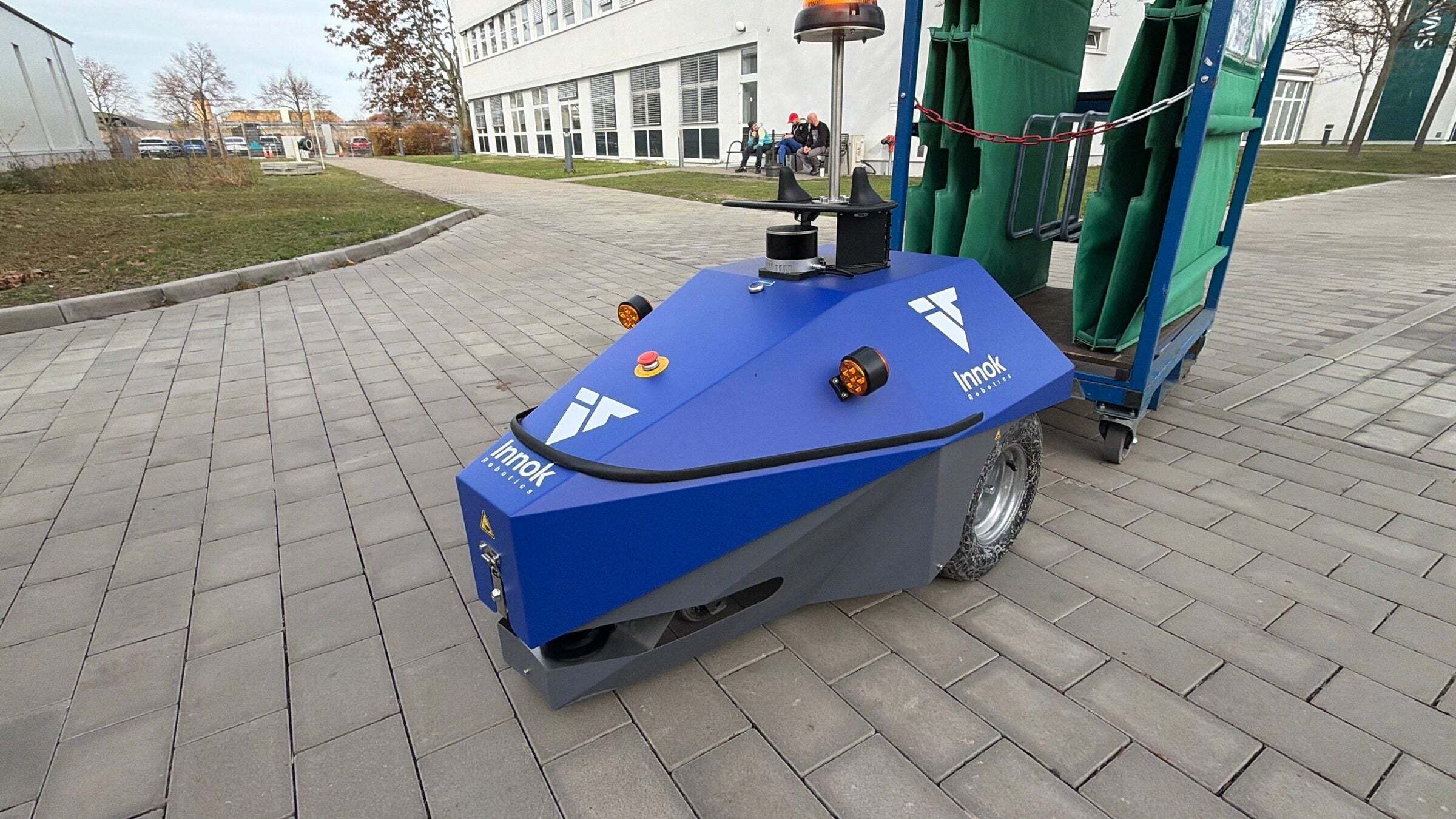 INDUROS autonomous mobile robot towing a trolley with green containers on a paved path outdoors at a factory site