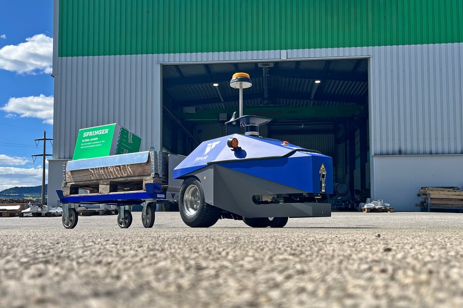 Blauer Transportroboter INDUROS mit Warnleuchte zieht einen Palettenwagen mit Palette über eine Außenfläche vor einer offenen Werkshalle