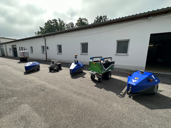 Mehrere Transportroboter INDUROS, die Roboterplattform HEROS und der Gießroboter RAINOS stehen vor einem Gebäude Spalier